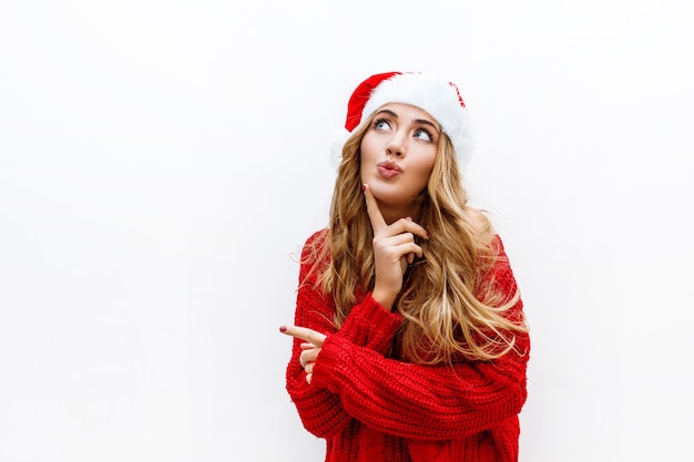 Blije onbezorgde blonde vrouw in nieuwe jaarhoed in het rode gebreide sweater stellen op witte muur. Isoleren. Kerst- en nieuwjaarsfeestconcept.