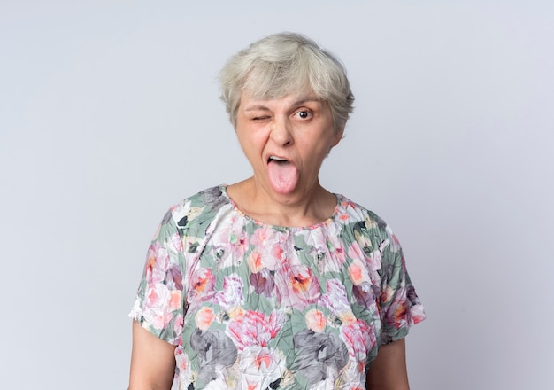 Blije oudere vrouw knippert in het oog en steekt tong uit die op witte muur wordt geïsoleerd