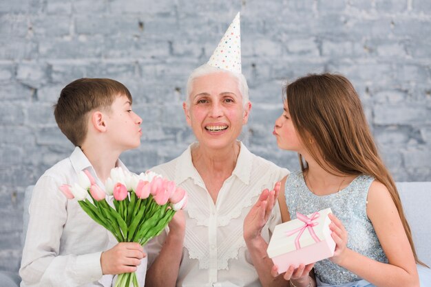Blije oudere vrouwenzitting binnen - tussen haar kleinkinderen met bloemenboeket en giftdoos