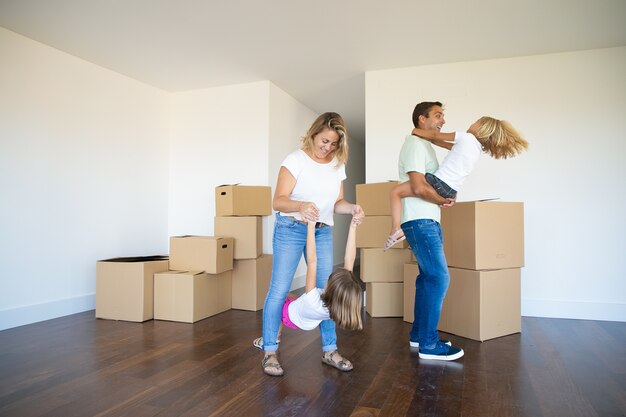 Blije ouders en kinderen die genieten van een nieuw huis, dansen en plezier maken in de buurt van hopen dozen in de lege ruimte