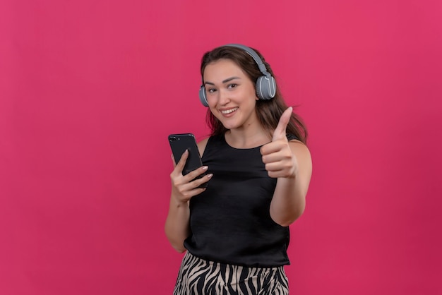 Blije vrouw die zwart hemd draagt, luistert muziek van hoofdtelefoons en haar duim omhoog op roze muur