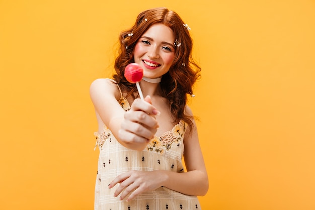 Blije vrouw in gele zomerjurk houdt roze snoep. Portret van vrouw met bloemen in haar haar op oranje achtergrond.