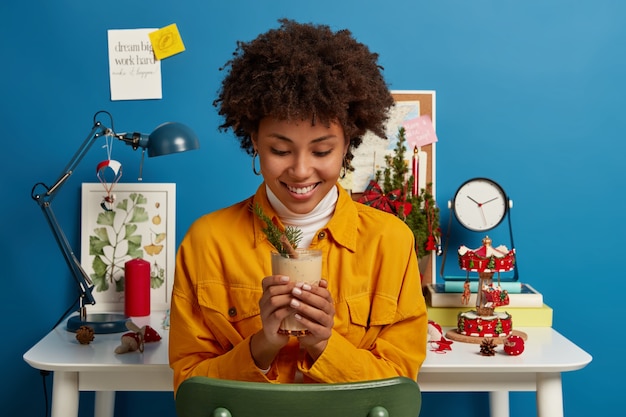 Blije vrouw met donkere huid die graag advocaatcocktail proeft, lacht aangenaam, wacht op kerstvakantie, poseert op stoel bij wit bureau met lamp, horloge, versierde dennenboom, heeft een feestelijke stemming