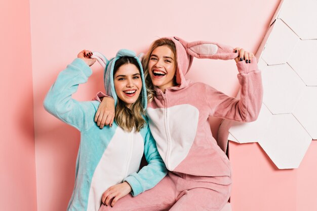 Blije zusters in kigurumi die zich op roze muur bevinden