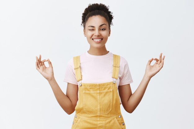 Blijf kalm en vrij van stress met meditatie. Vrolijke en speelse stijlvolle moderne Afro-Amerikaanse vrouw in gele overall, gluren met één oog en breed glimlachend, hand in hand in zen pose