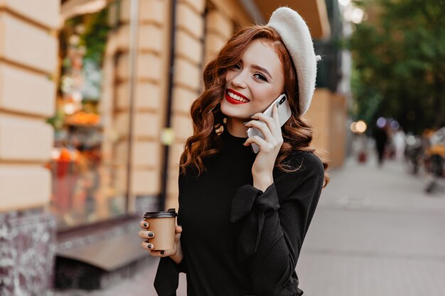 Blithesome europees meisje dat in baret op stadsmuur glimlacht. Glamoureuze langharige dame praten aan de telefoon en koffie drinken.