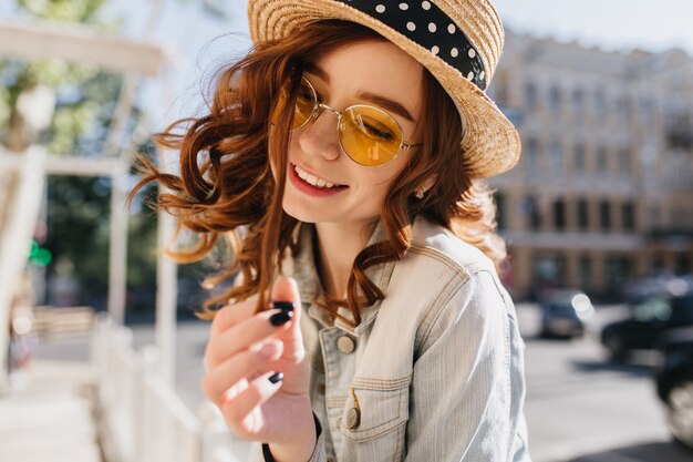 Blithesome europees meisje speelt met haar gemberhaar met een glimlach. Buiten schot van gelukkig roodharige dame in zomer hoed poseren op straat.