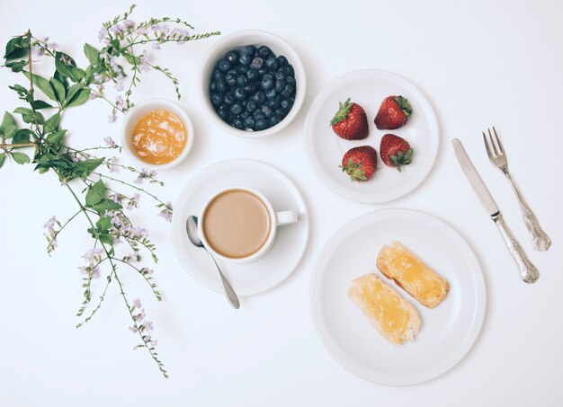 Bloem; jam; bosbes; aardbei; koffiekopje en toast brood op witte achtergrond met bestek