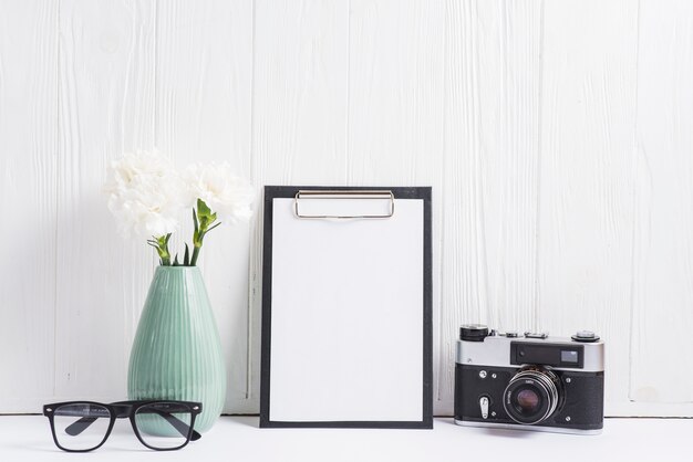 Bloemenvaas; bril; camera en blanco papier op klembord tegen lege houten muur