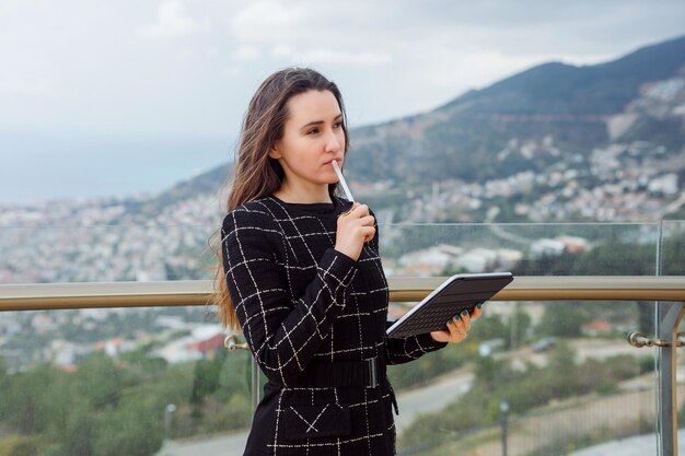 Blogger-meisje denkt door de pen op de mond te houden tegen de achtergrond van het uitzicht op de stad