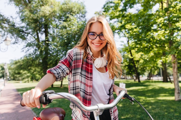 Blond blij meisje dat rond park in ochtend berijdt. Buitenfoto van betoverende jonge dame met fiets die positieve emoties uitdrukt.