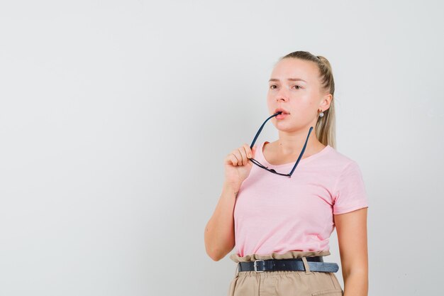 Blond meisje bijt bril in t-shirt, broek en op zoek doordachte, vooraanzicht.