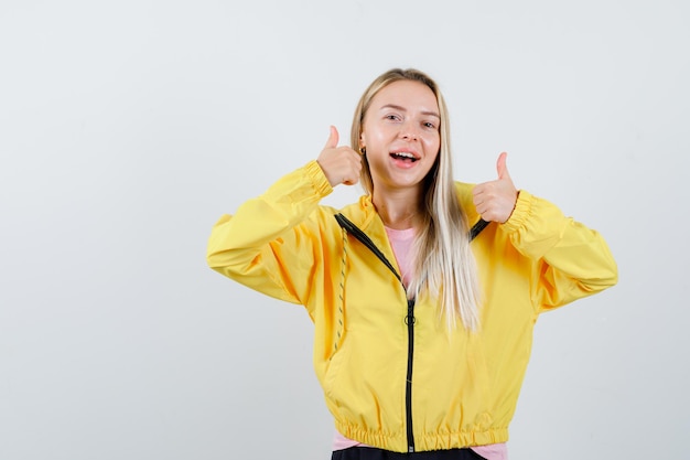 Blond meisje dat dubbele duimen in gele jas laat zien en er gelukkig uitziet