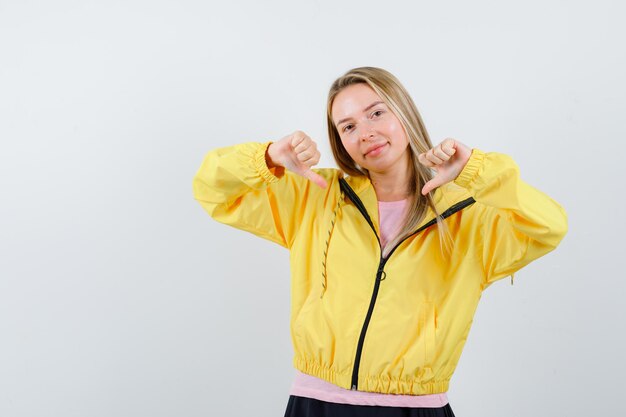 Blond meisje dat duimen naar beneden laat zien met beide handen in roze t-shirt en geel jasje en er gelukkig uitziet