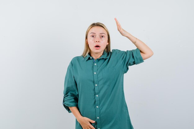 Blond meisje dat haar hand boven het hoofd houdt terwijl ze haar hand op de buik houdt in een groene blouse en er verbaasd uitziet