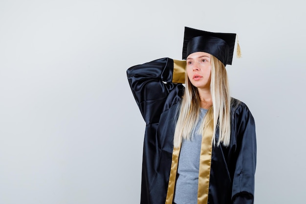 Gratis foto blond meisje dat hand achter haar hoofd zet, aan iets denkt in afstudeerjurk en pet en peinzend kijkt