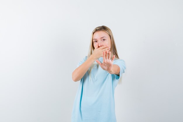 Blond meisje dat mond bedekt met hand, stopbord in blauw t-shirt toont en verbaasd, vooraanzicht kijkt.