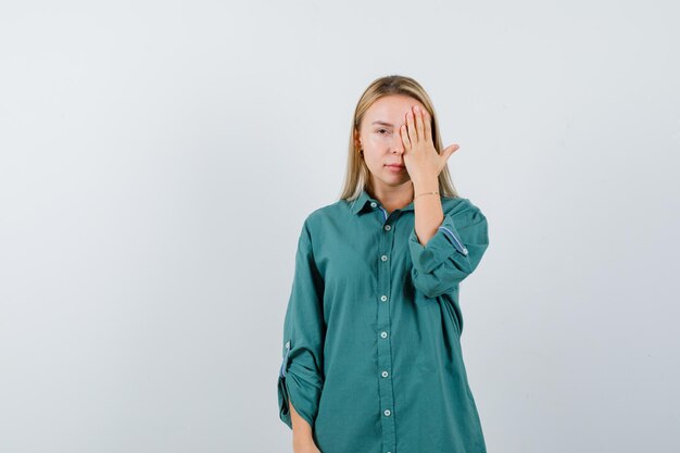 Blond meisje dat oog bedekt met hand in groene blouse en er serieus uitziet