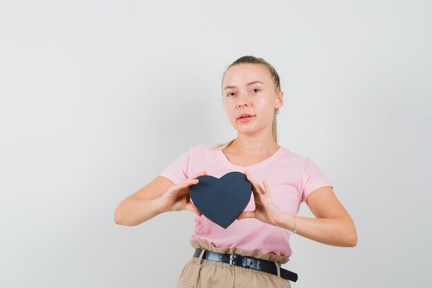 Blond meisje geschenkdoos in t-shirt, broek houden en op zoek naar zelfverzekerd, vooraanzicht.