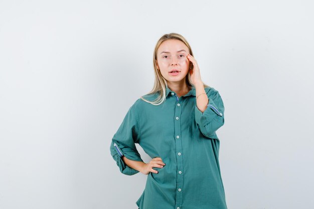 Blond meisje houdt de hand op de taille terwijl ze de hand op de wang legt in een groene blouse en er serieus uitziet.