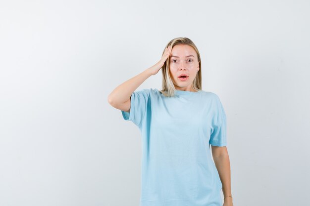 Blond meisje houdt de hand op tempel in blauw t-shirt en kijkt verbaasd. vooraanzicht.