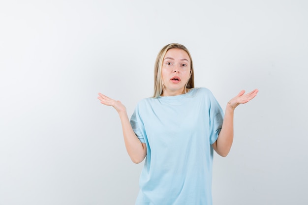 Blond meisje hulpeloos gebaar in blauw t-shirt tonen en op zoek verward. vooraanzicht.