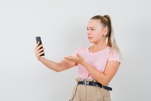 Blond meisje iets bespreken op videochat in t-shirt, broek en op zoek verward, vooraanzicht.