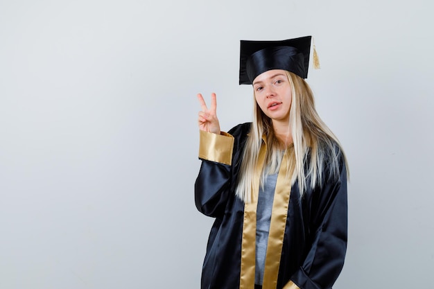 Gratis foto blond meisje in afstudeerjurk en pet die vredesgebaar toont en er schattig uitziet