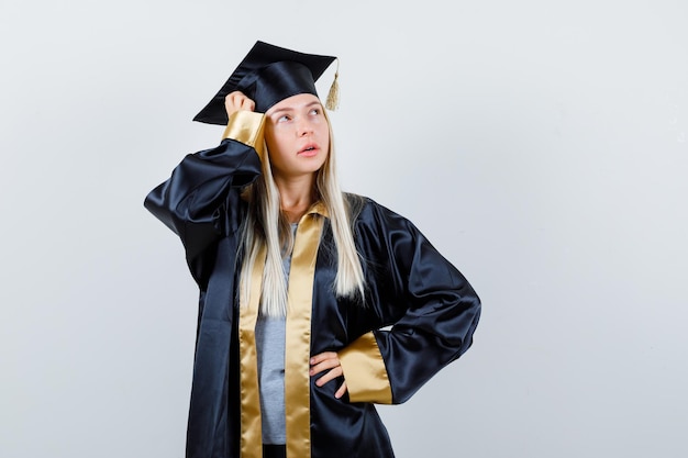 Blond meisje in afstudeerjurk en pet met hand op taille, hand op hoofd, wegkijkend en zelfverzekerd kijkend