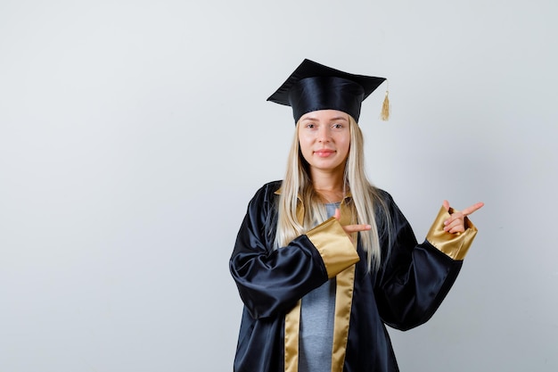 Blond meisje in afstudeerjurk en pet wijst naar rechts met wijsvingers en ziet er schattig uit