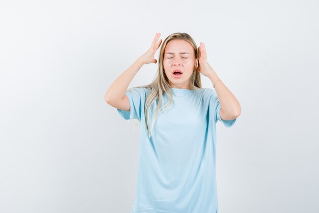 Blond meisje in blauw t-shirt hand in hand in de buurt van hoofd en op zoek geërgerd, vooraanzicht.