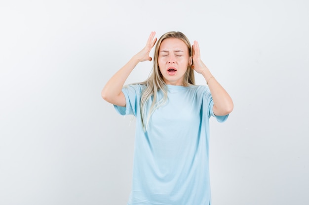 Blond meisje in blauw t-shirt hand in hand in de buurt van hoofd en op zoek geërgerd, vooraanzicht.