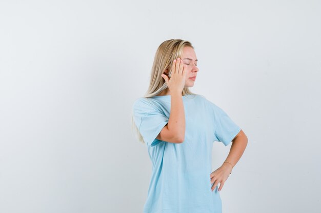 Blond meisje in blauw t-shirt hand op de tempel zetten, hand op de taille te houden en op zoek moe, vooraanzicht.