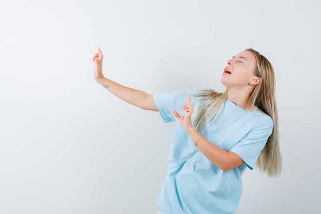 Blond meisje in blauw t-shirt met geweergebaren, ogen gesloten houden en op zoek vrolijk, vooraanzicht.