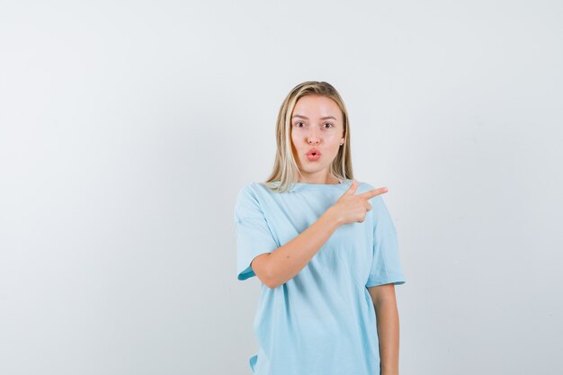 Blond meisje in blauw t-shirt naar rechts met wijsvinger en op zoek verrast, vooraanzicht.