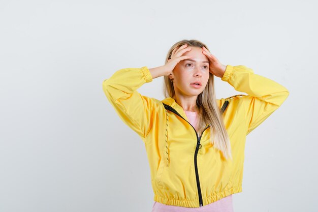 Blond meisje in gele jas wrijft over haar voorhoofd en kijkt bedachtzaam