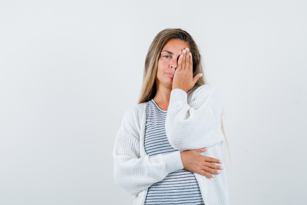 Blond meisje in gestreept t-shirt, wit vest en jean broek die oog met hand bedekt en verleidelijk, vooraanzicht kijkt.