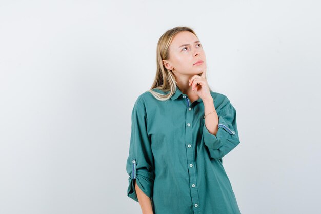 Blond meisje in groene blouse staat in denkende pose en kijkt peinzend