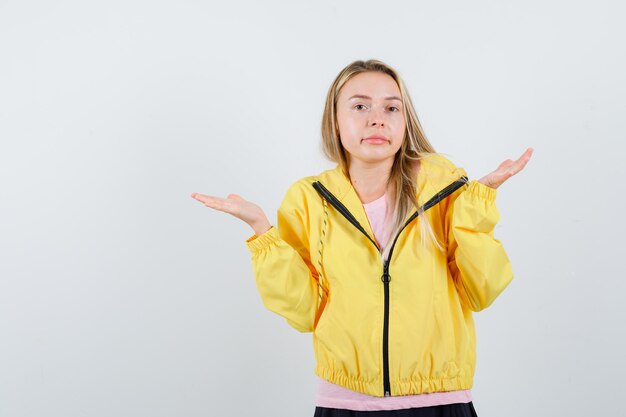 Blond meisje in roze t-shirt en gele jas die handen op een vragende manier uitrekt en verbaasd kijkt