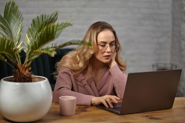 Blond meisje in roze vest en bril. werken op haar laptop. zittend aan houten tafel met roze kop, palm in pot erop. student, blogger. interieur met witte bakstenen muur en groene bank. detailopname
