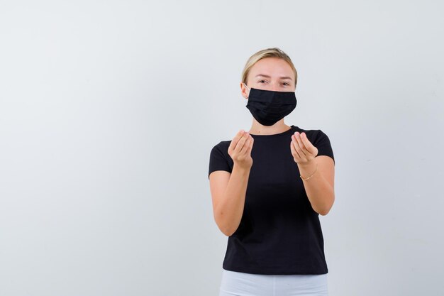 Blond meisje in zwart t-shirt, witte broek, zwart masker met Italiaans gebaar geïsoleerd