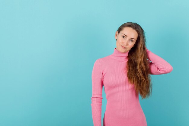 Blond meisje kijkt naar de camera door de hand achter het hoofd op een blauwe achtergrond te houden