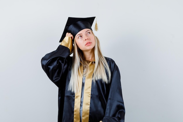 Gratis foto blond meisje krabt aan het hoofd, denkt aan iets in afstudeerjurk en pet en kijkt peinzend.