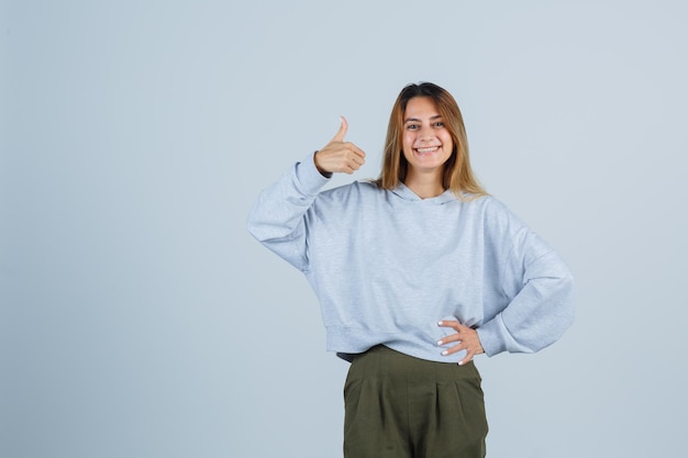 Blond meisje met duim omhoog en hand op taille in olijfgroen blauw sweatshirt en broek en ziet er gelukkig uit. vooraanzicht.