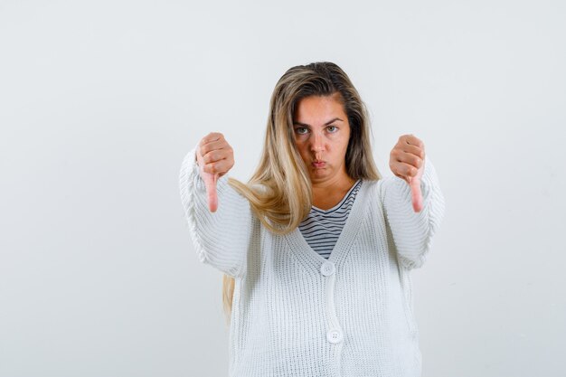 Blond meisje met duimen naar beneden met beide handen in gestreept t-shirt, wit vest en jean broek en op zoek naar ontevreden, vooraanzicht.