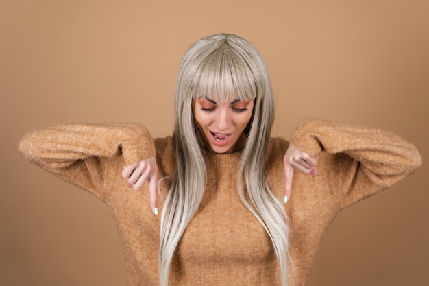 Gratis foto blond meisje met pony en bruine make-up voor overdag in een trui op een beige achtergrond glimlacht opgewonden en wijst haar vinger naar beneden