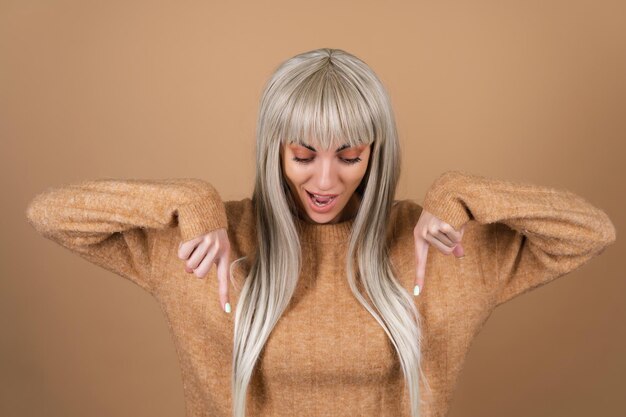 Blond meisje met pony en bruine make-up voor overdag in een trui op een beige achtergrond glimlacht opgewonden en wijst haar vinger naar beneden