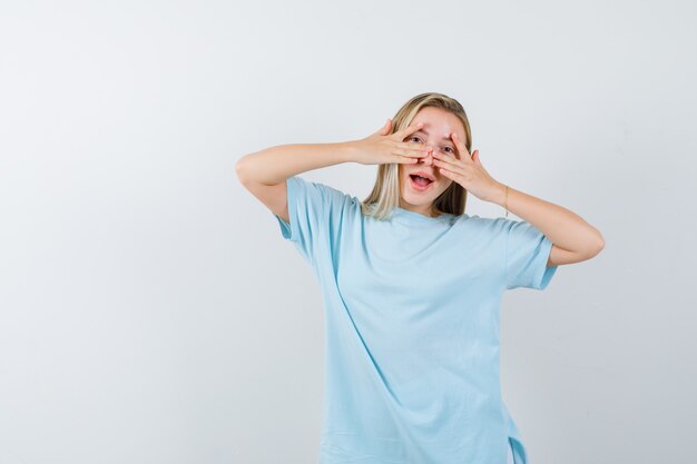 Blond meisje met v-tekens op ogen in blauw t-shirt en ziet er mooi uit, vooraanzicht.