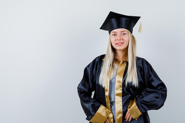 Gratis foto blond meisje staat rechtop, glimlacht en poseert voor de camera in afstudeerjurk en pet en ziet er schattig uit.