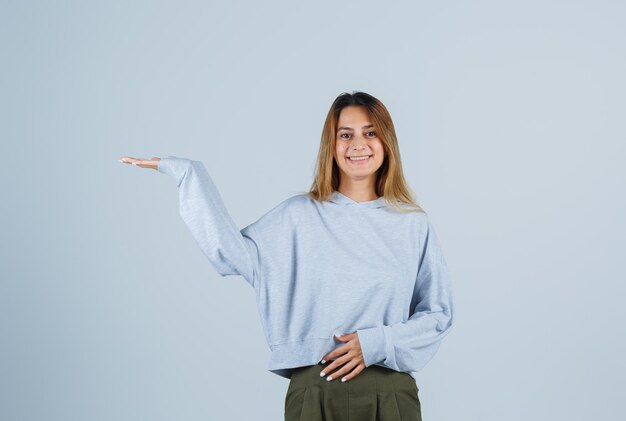 Blond meisje strekt haar handen uit alsof ze iets vasthoudt terwijl ze een hand op de buik houdt in een olijfgroen blauw sweatshirt en broek en er gelukkig uitziet. vooraanzicht.